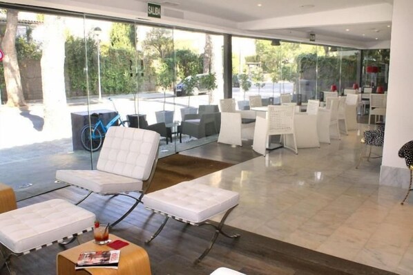 A modern indoor seating area with white cushioned chairs and tables, large windows offering a view of the outdoors, and a sign indicating the exit. - Sitges (Sitges)