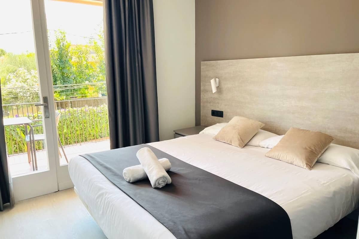Habitación de hotel con una cama grande, una mesita de noche, una lámpara de pared y vistas al exterior a través de una puerta corrediza.