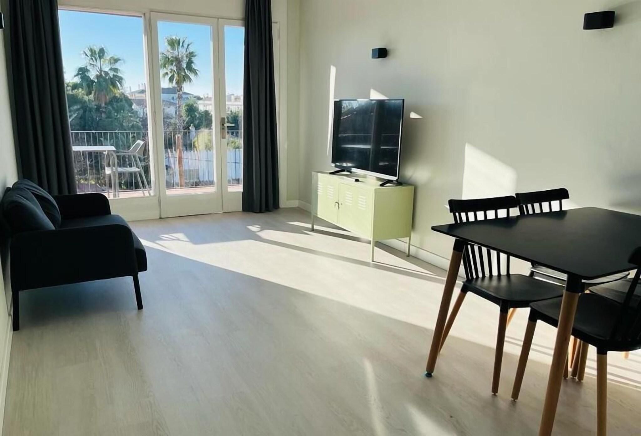 A modern living room with a sofa, a dining table with chairs, a TV on a cabinet, and a balcony with a view of palm trees and buildings.