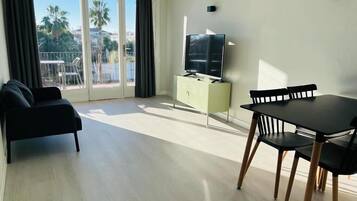 A modern living room with a sofa, a dining table with chairs, a TV on a cabinet, and a balcony with a view of palm trees and buildings.