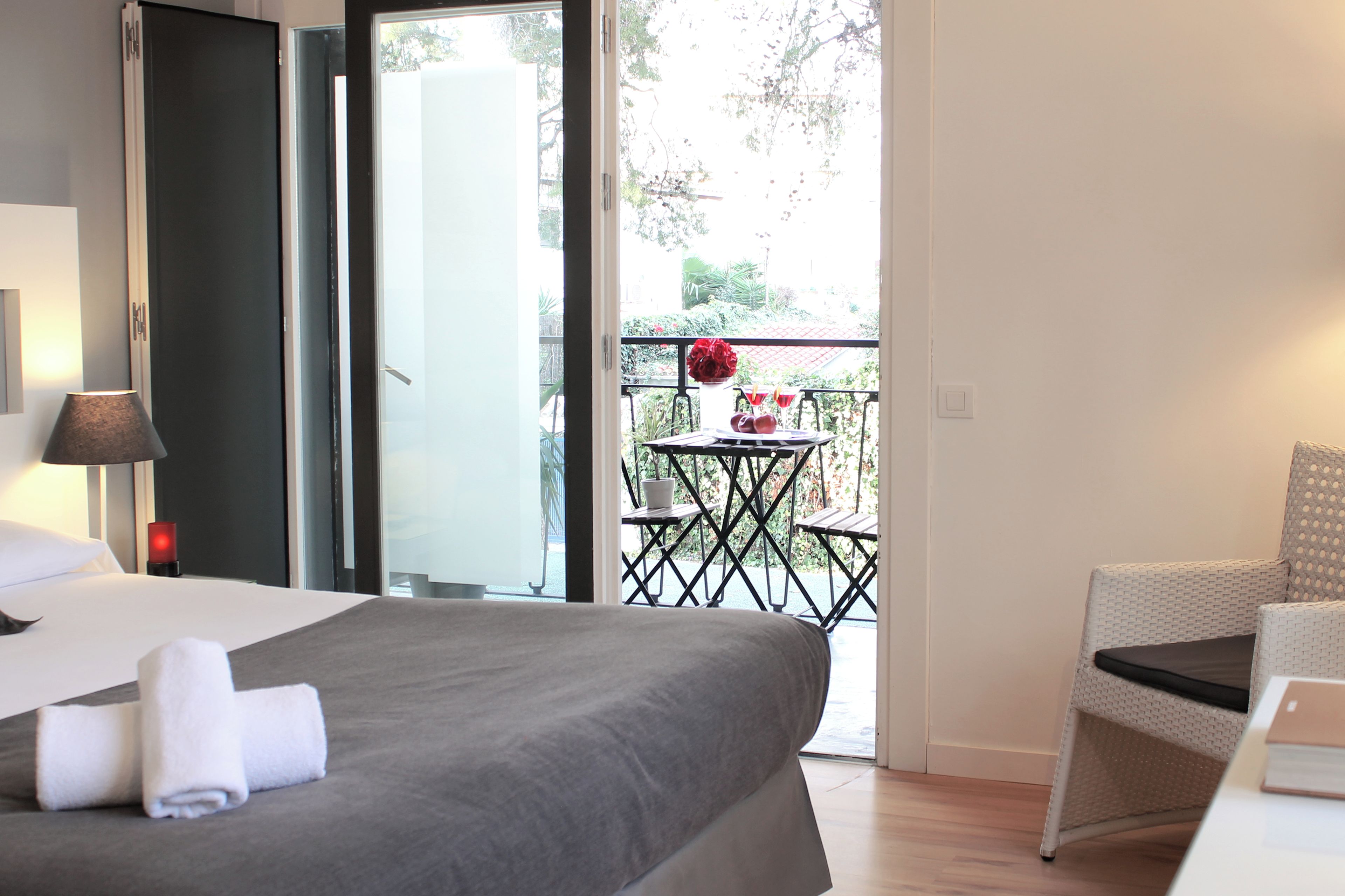 Una habitación de hotel con cama, una mesa con un jarrón de flores rojas y un balcón con mesa y sillas.