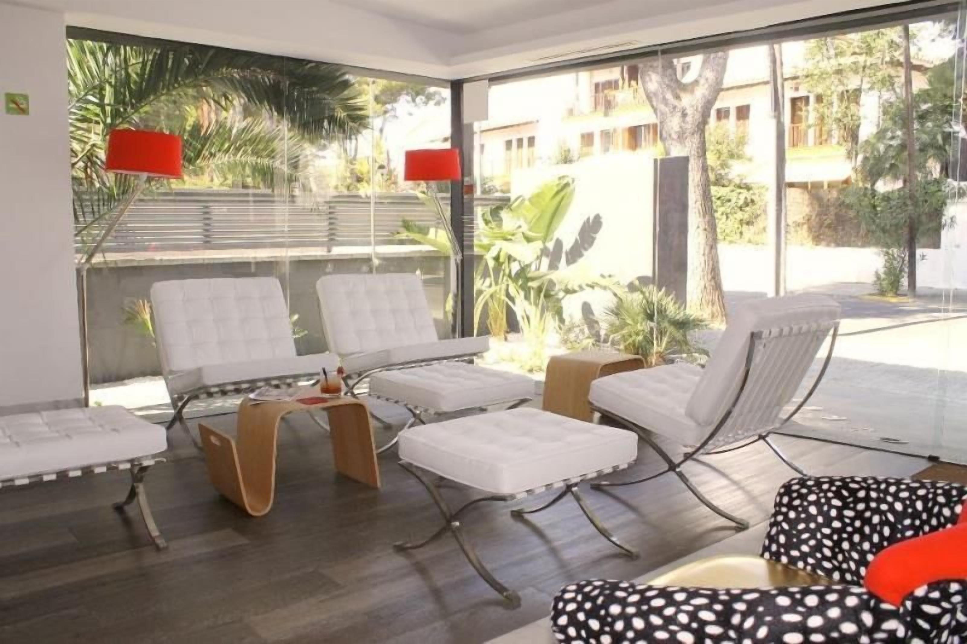A modern hotel room with a large window, white cushioned chairs, a small wooden table, and a red lamp.