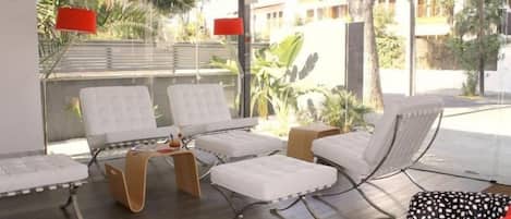 A modern hotel room with a large window, white cushioned chairs, a small wooden table, and a red lamp.