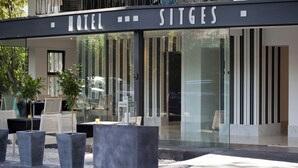A modern hotel entrance with a glass facade, outdoor seating, and potted plants. - Sitges (Sitges)