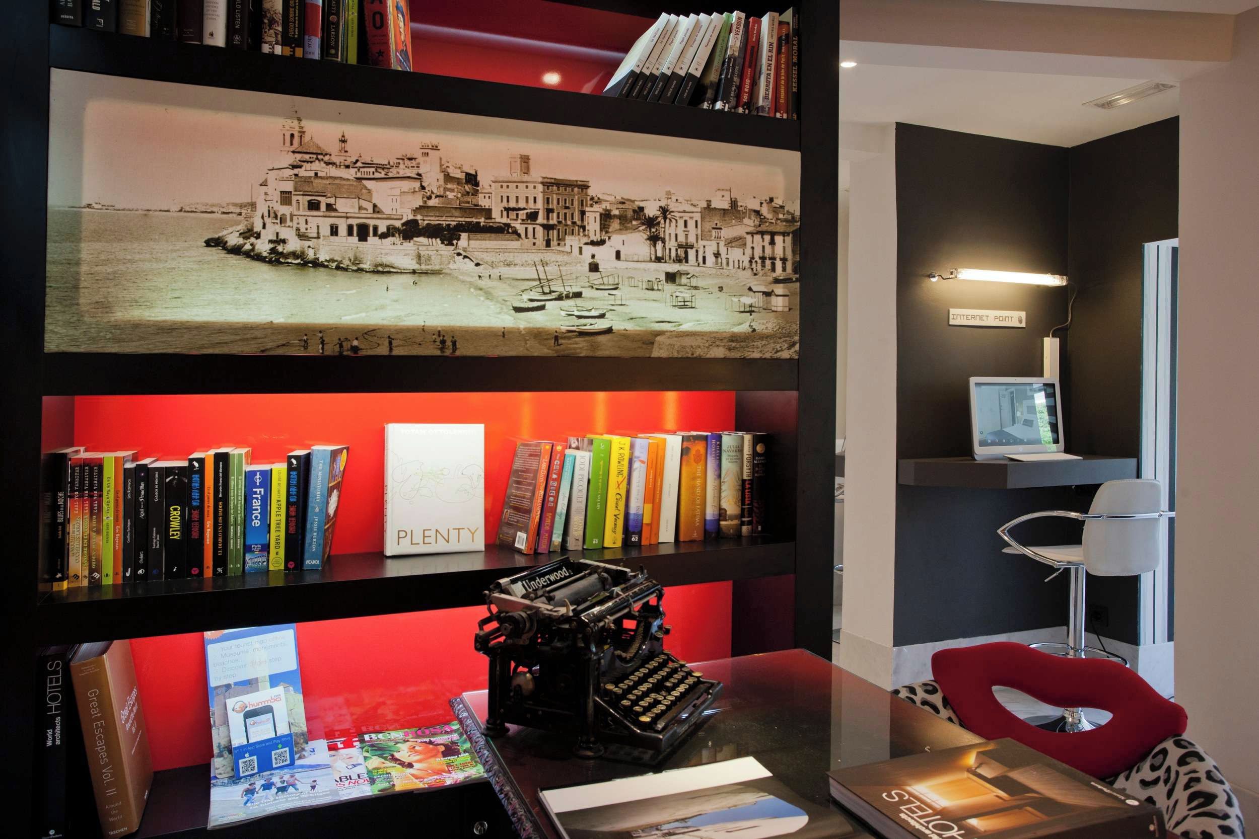Una habitación con una estantería llena de libros y una máquina de escribir, un escritorio con un monitor de computadora, y una fotografía histórica enmarcada.