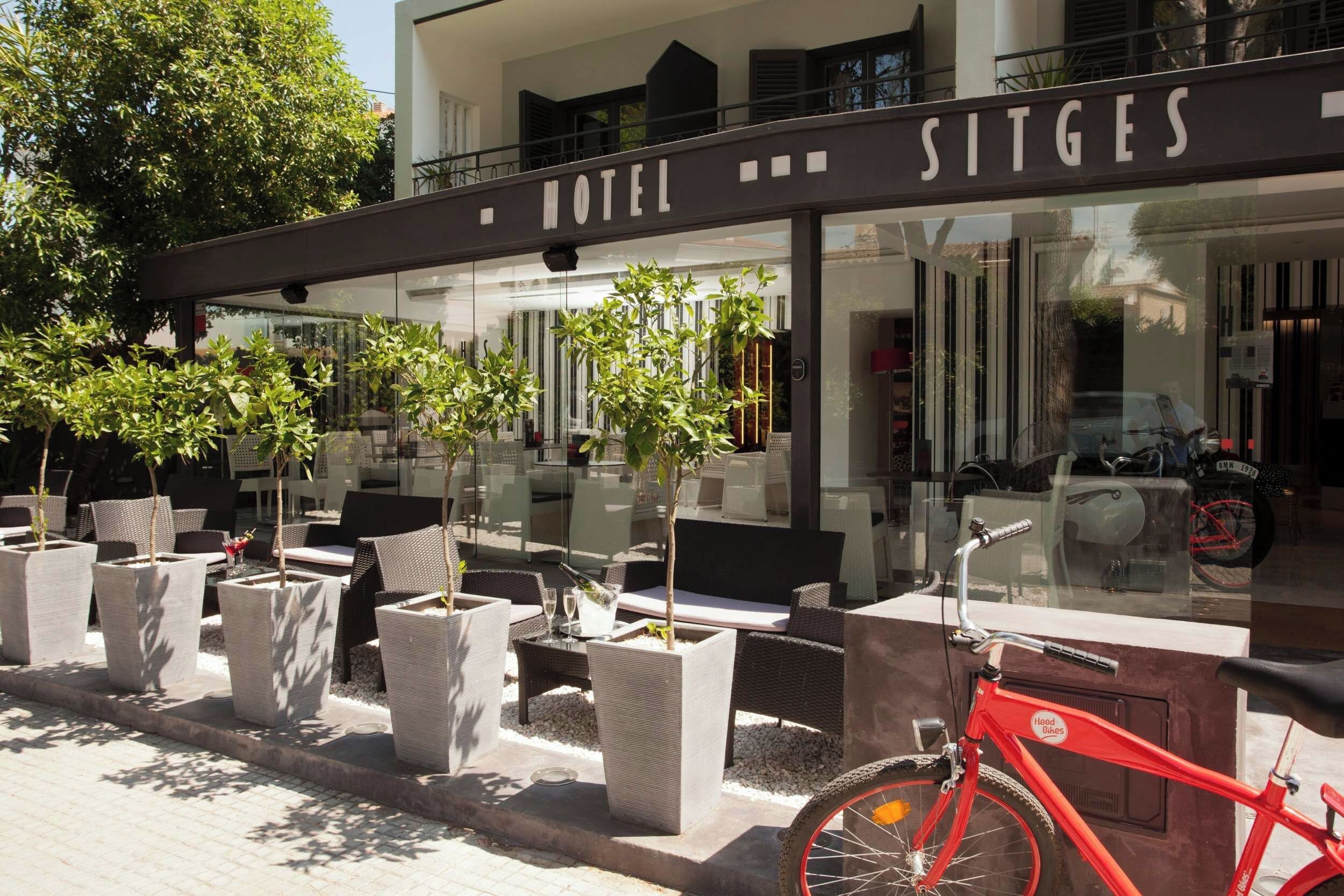 Exterior de un hotel con mesas al aire libre, plantas en macetas y una bicicleta roja.