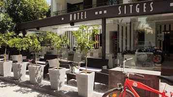 Exterior de un hotel con mesas al aire libre, plantas en macetas y una bicicleta roja.