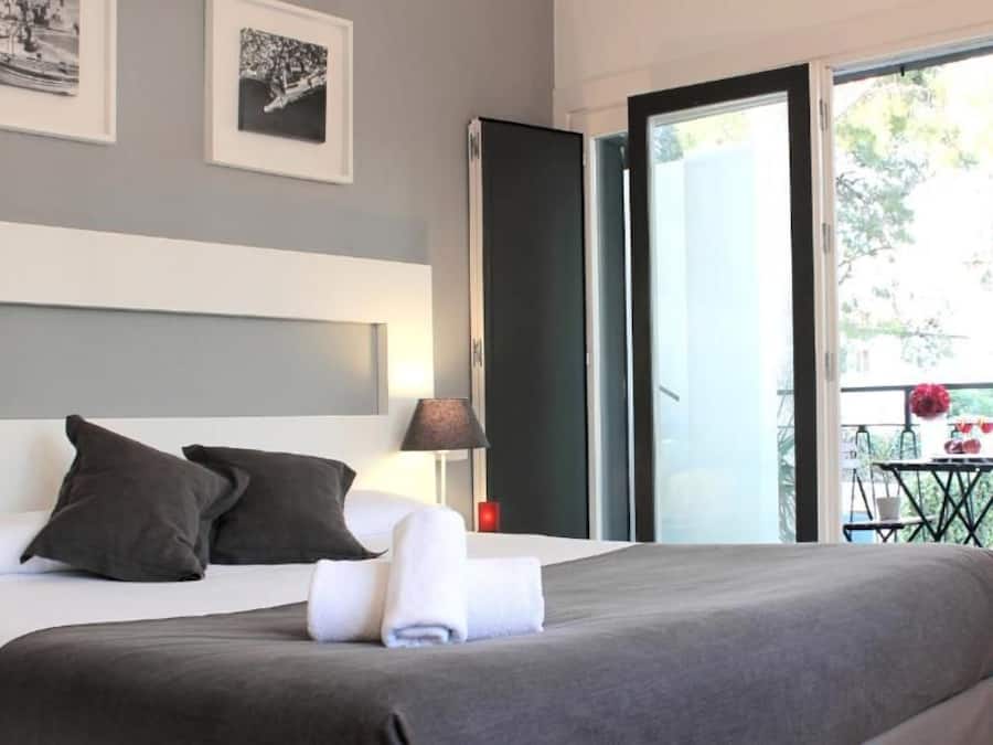 Una habitación de hotel moderna con una cama grande, una mesita de noche, una lámpara y un balcón con mesa y sillas.