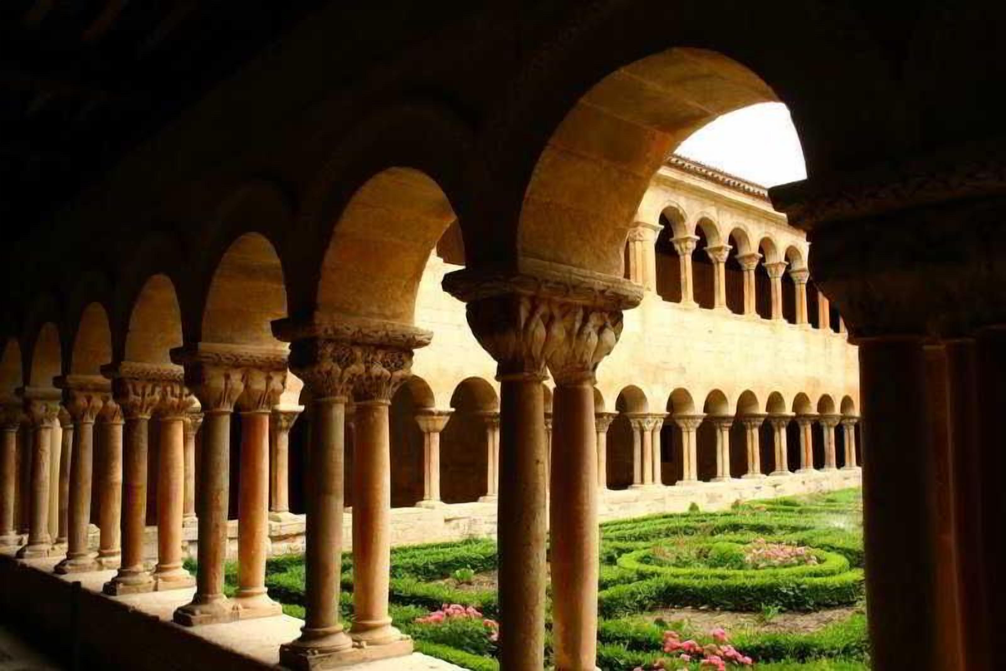 Una serie de arcos con columnas, un jardín con setos podados y un edificio con fachada de tonos claros.