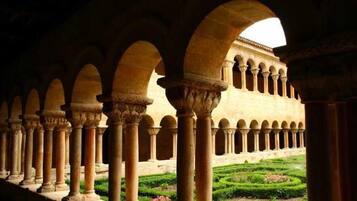 Una serie de arcos con columnas, un jardín con setos podados y un edificio con fachada de tonos claros.