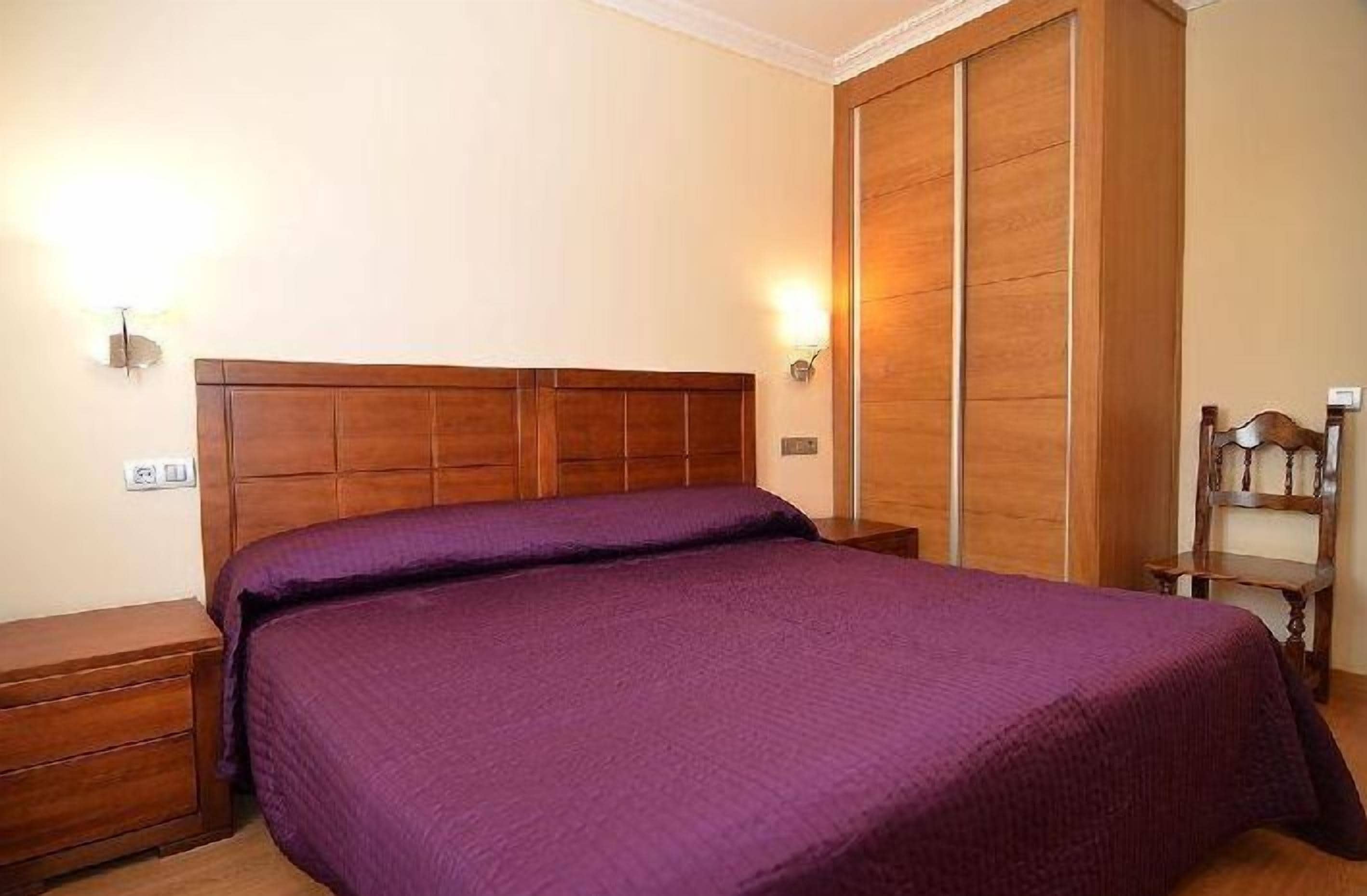 Un dormitorio con cabecera de madera, colcha morada, mesita de noche, armario de madera y una pequeña silla de madera.