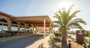 A hotel entrance with a covered walkway, palm trees, and a clear sky. - Ilunion Sancti Petri (Chiclana de la Frontera)
