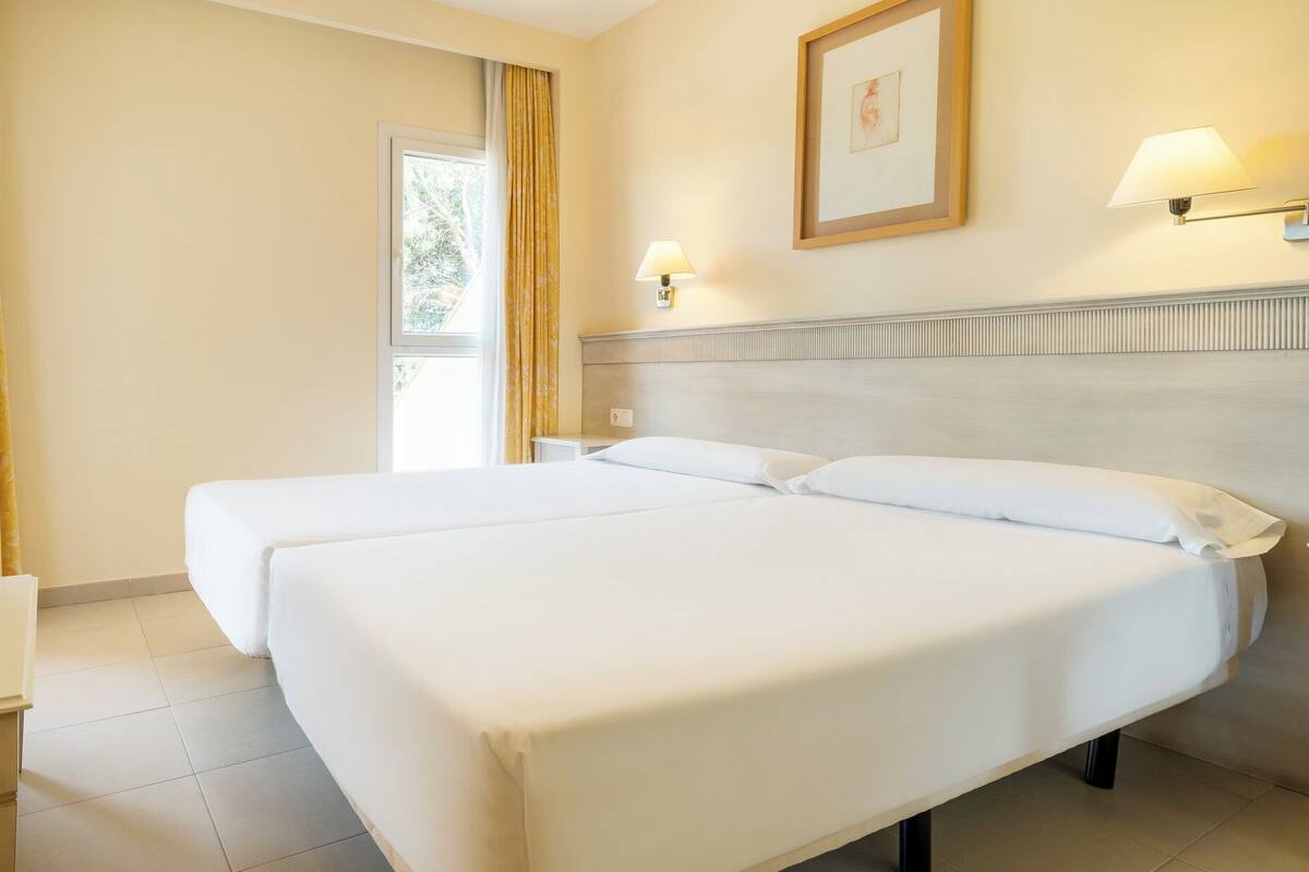 A hotel room with two beds, a small table, and a window with curtains.