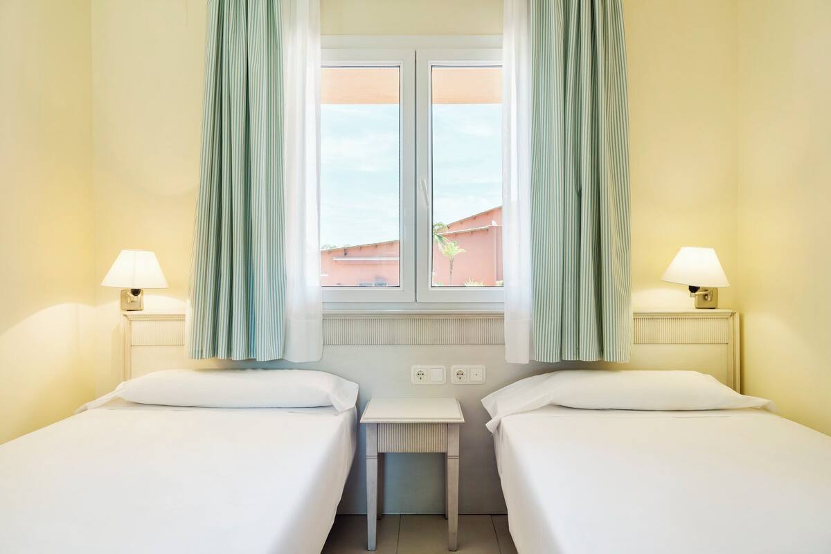 A hotel room with two single beds, a small table, two bedside lamps, and a window with curtains.