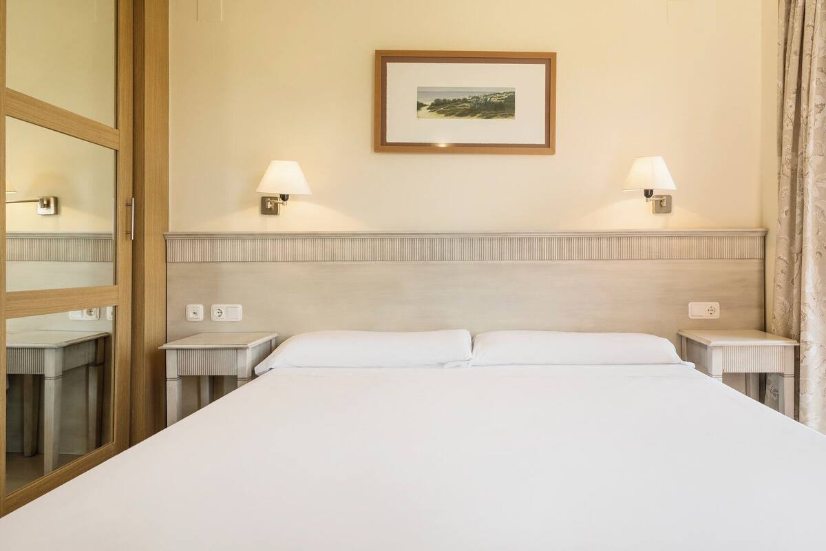 A hotel room with a large bed, two bedside tables, two wall-mounted lamps, a framed picture on the wall, and a window with curtains.