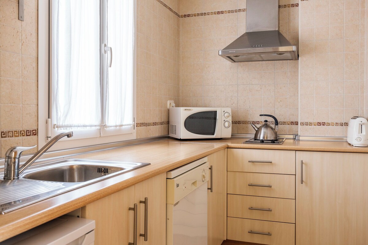 A kitchen with a sink, a microwave, a kettle, and wooden cabinets.