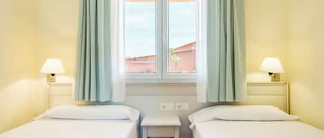 A hotel room with two single beds, a small table, two bedside lamps, and a window with curtains.