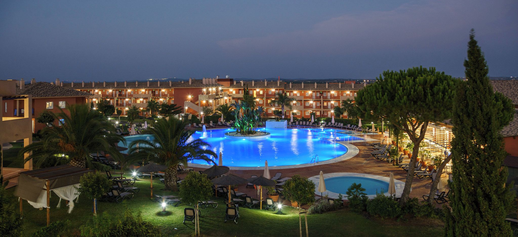 Un hotel con una amplia piscina exterior, varias zonas de descanso y un jardín bien iluminado.