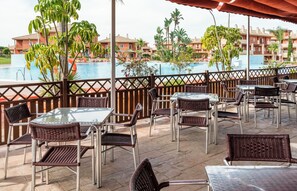 A poolside patio with wicker chairs and tables, overlooking a pool and a building complex. - Ilunion Sancti Petri (Chiclana de la Frontera)