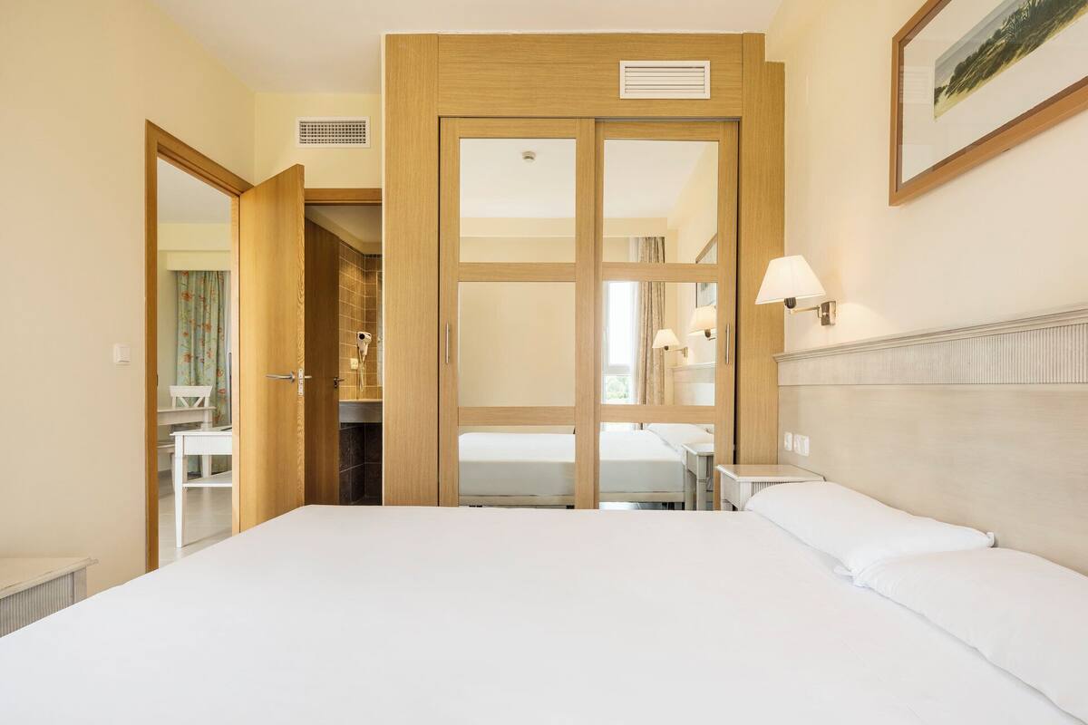 A hotel room with a large bed, a wooden wardrobe, a bathroom with a shower, and a small table with a lamp.