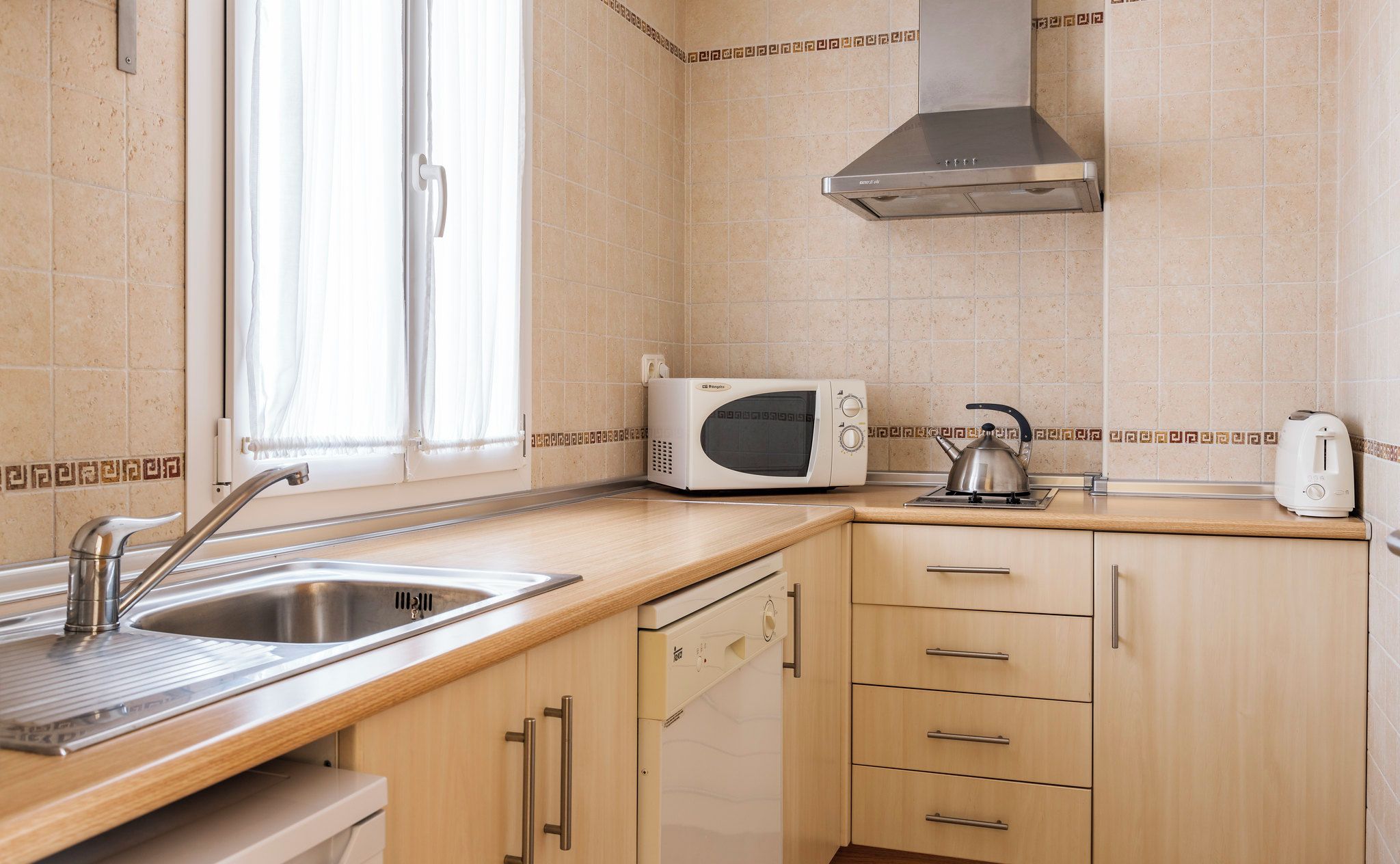 A kitchen with a stainless steel sink, a microwave, a kettle, and wooden cabinets.