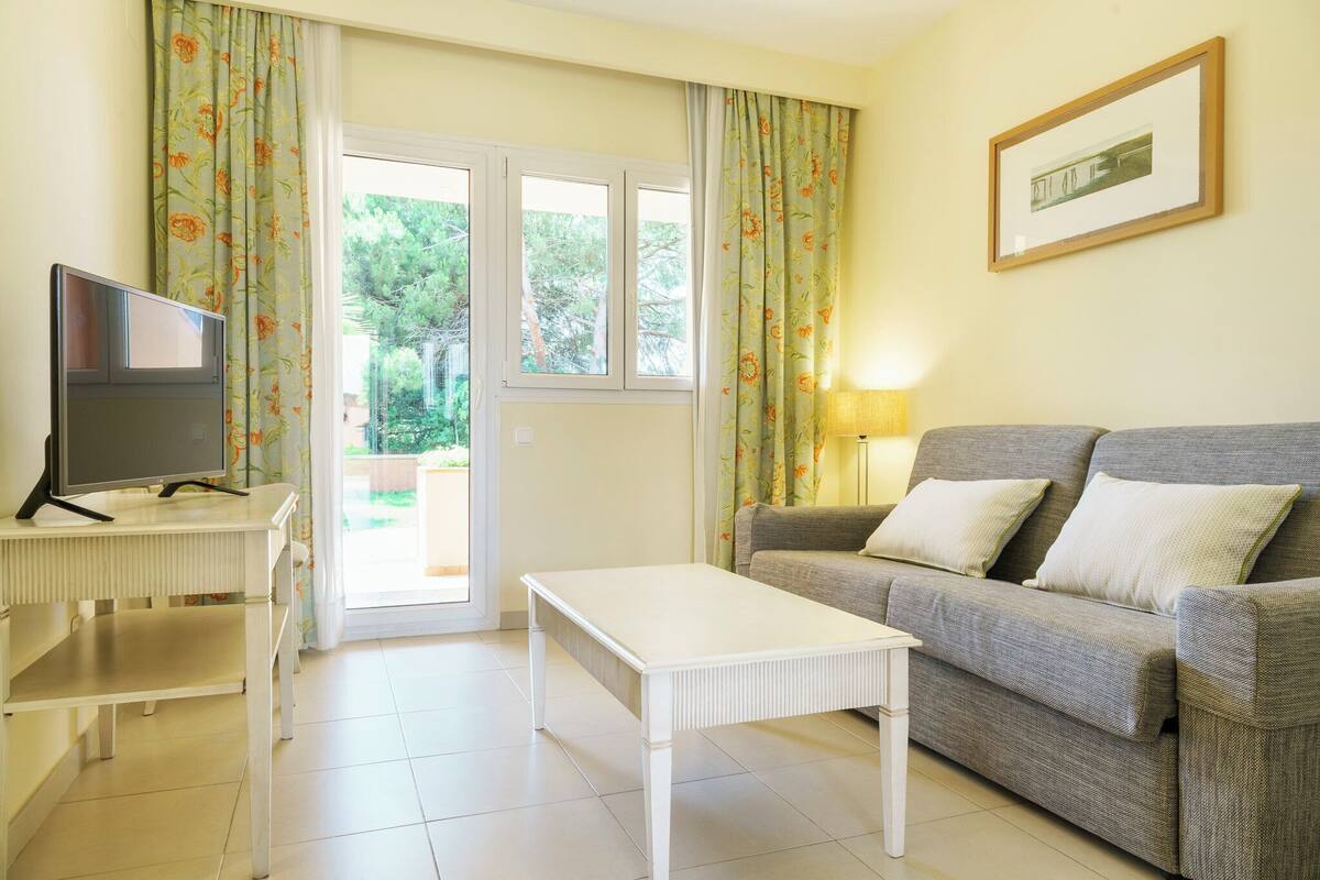 A living room with a sofa, a TV on a stand, a table, and a door leading outside.