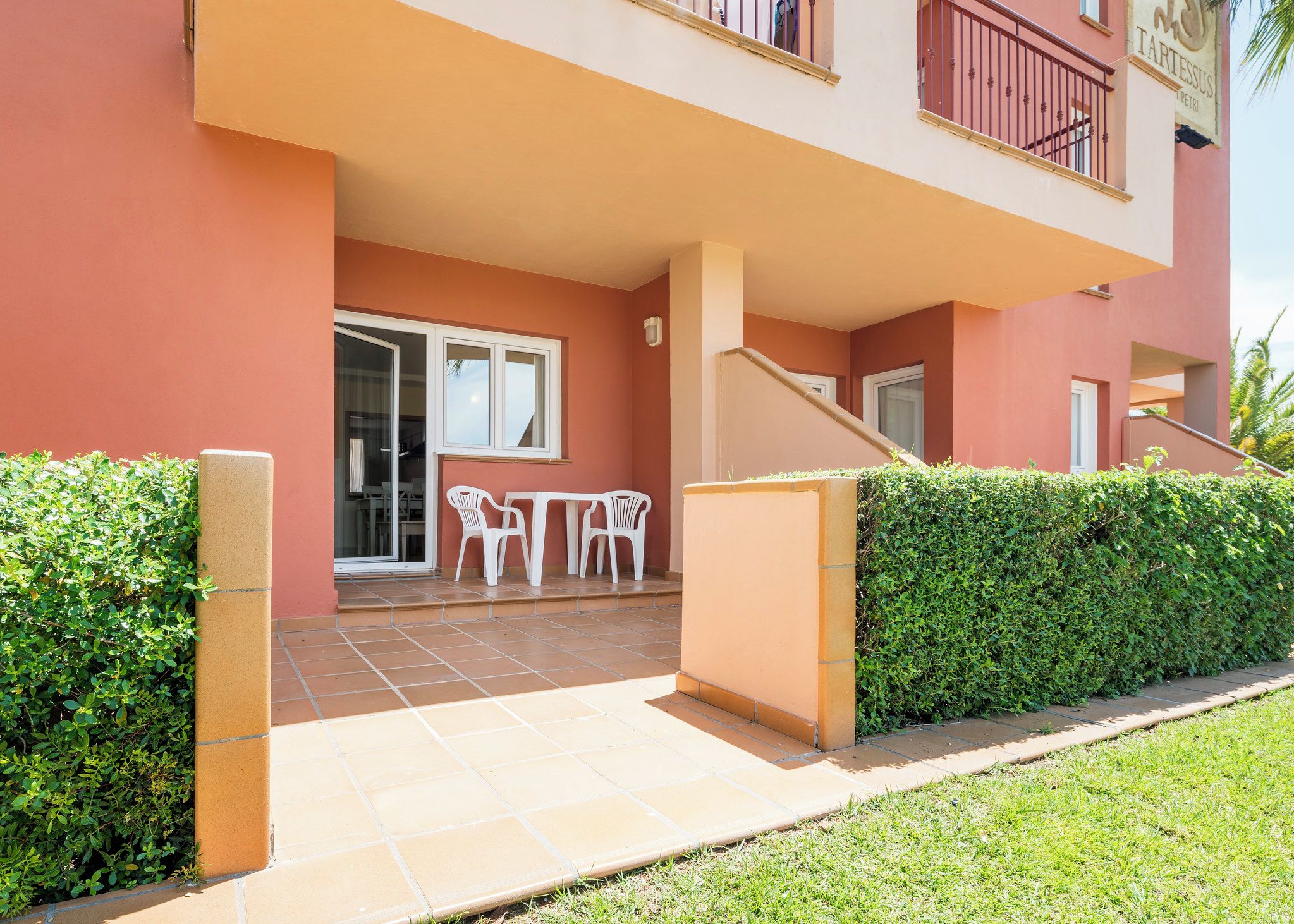 Un bâtiment à l’extérieur rouge, doté d’un patio couvert avec des chaises et une table en plastique blanc, et d’un escalier menant à un étage supérieur.