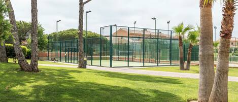 A tennis court enclosed by a green fence, surrounded by trees and a bench.