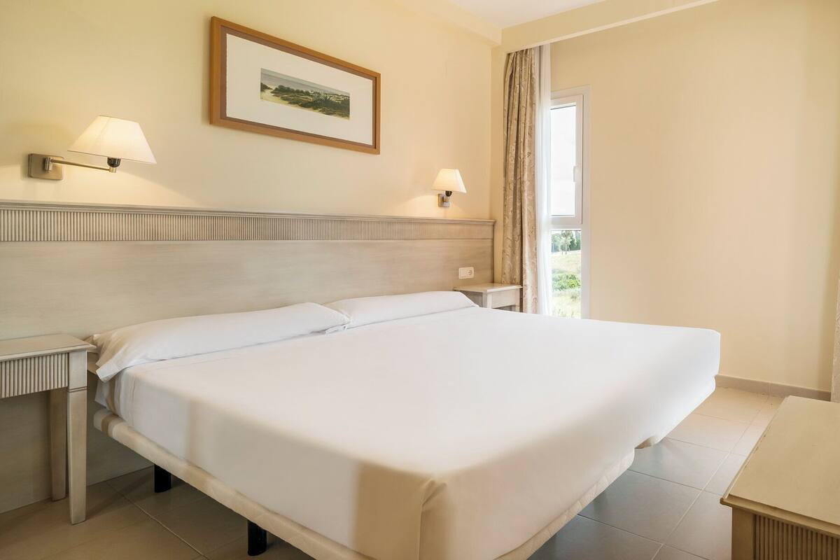 A hotel room with a large bed, two bedside lamps, a framed picture on the wall, and a window with curtains.