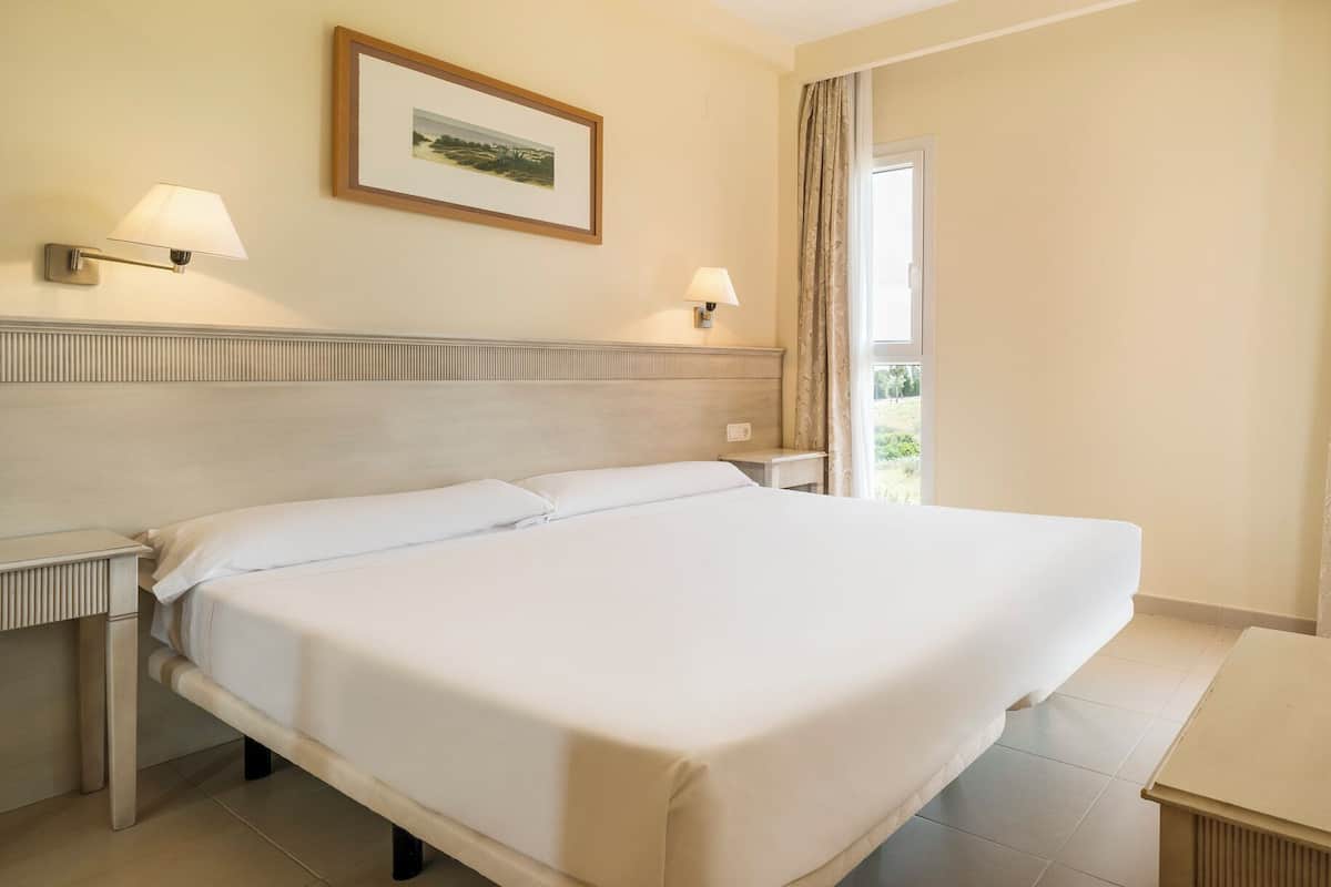 Habitación de hotel con una cama grande, dos lámparas de noche, un cuadro enmarcado en la pared y una ventana con cortinas.