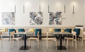 A modern dining area with tables and chairs, a sofa, and abstract leaf-patterned wall art. - Ilunion Sancti Petri (Chiclana de la Frontera)