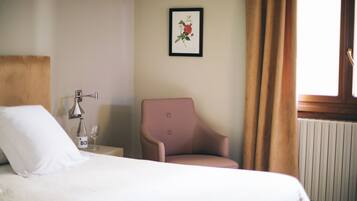 A hotel room with a bed, a chair, a bedside table with a bottle and glass, and a framed botanical print on the wall.