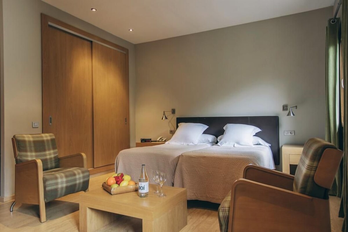 Une chambre d’hôtel avec un lit, deux chaises, une table basse en bois sur laquelle se trouvent des fruits et une bouteille, et une armoire.
