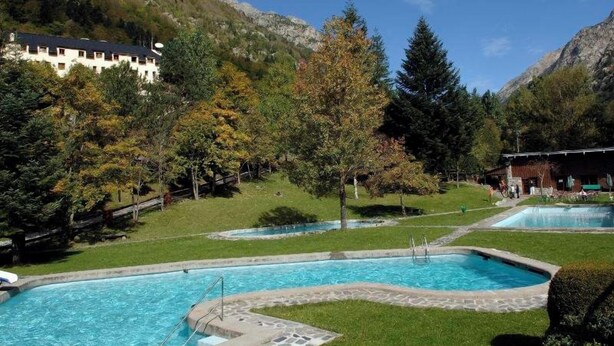 Un espace piscine comprenant plusieurs bassins, entouré d’arbres et offrant une vue sur un paysage montagneux.