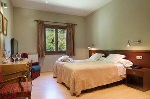 Habitación de hotel con dos camas, una mesita de noche de madera, un escritorio con silla, una ventana con cortinas y vistas a zonas verdes.