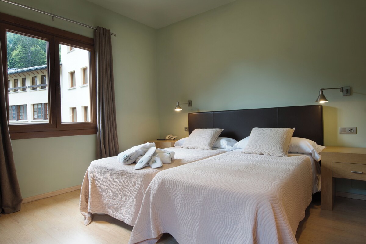 Une chambre d’hôtel avec deux lits, une table de chevet, une lampe et une fenêtre avec des rideaux.