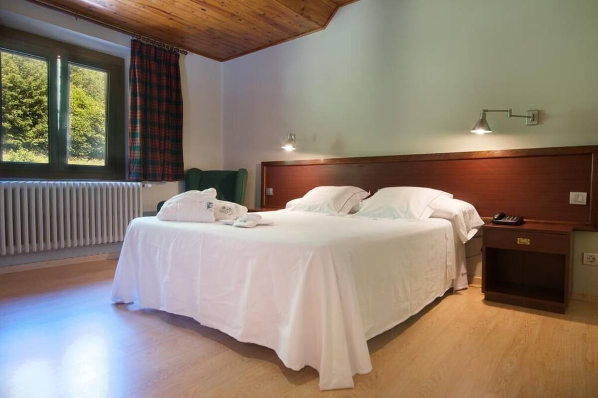 Un lit bien fait, avec du linge de lit blanc, une table de chevet avec une lampe, et une vue sur les arbres par la fenêtre.