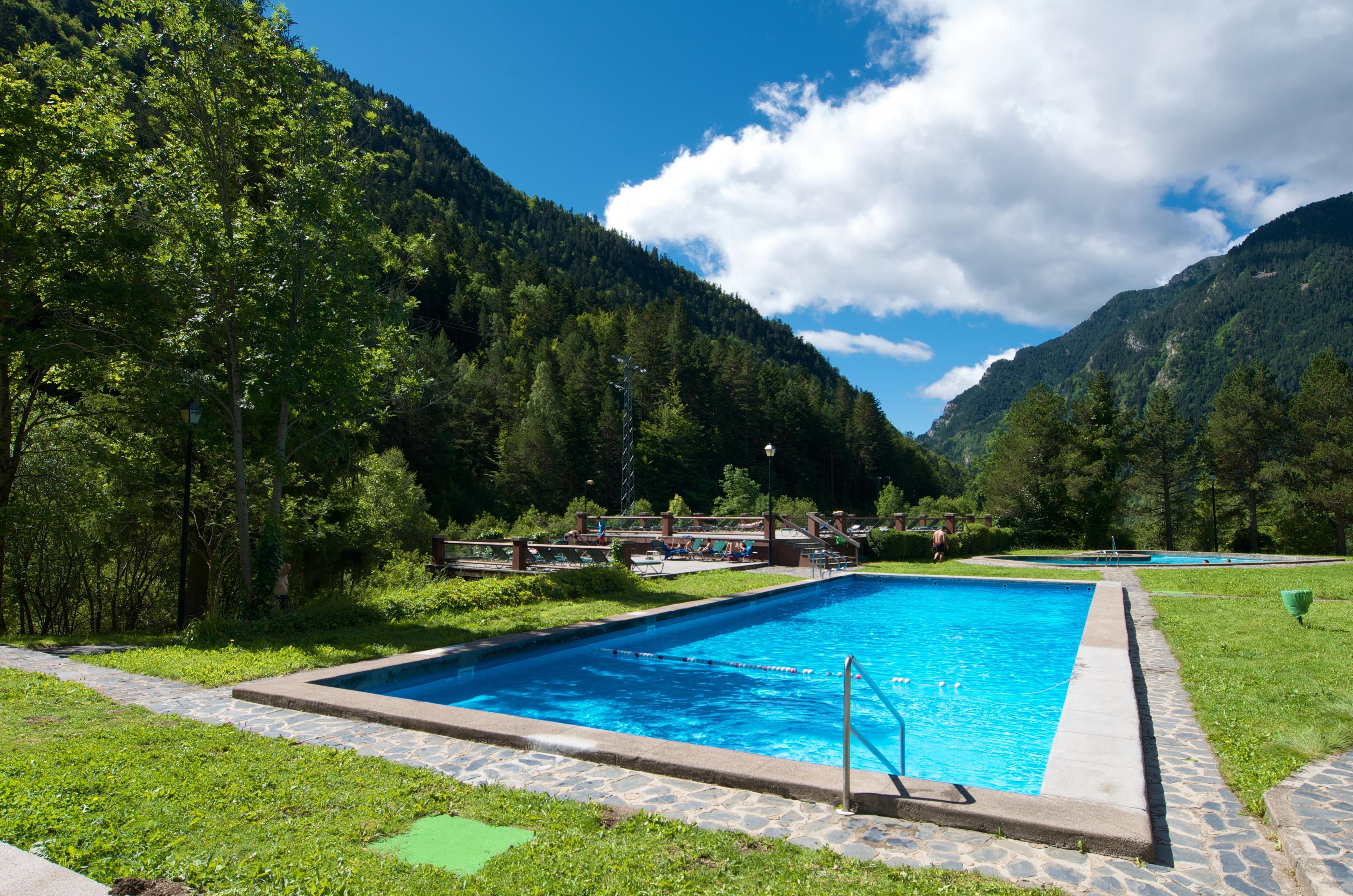 A rectangular swimming pool with a stone border, surrounded by a stone pathway, set against a backdrop of lush green trees and mountains under a partly cloudy sky.
