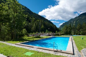 Une piscine rectangulaire bordée de pierres, entourée d’un sentier également en pierre, le tout se fondant dans un paysage verdoyant composé d’arbres luxuriants et de montagnes, sous un ciel partiellement nuageux.