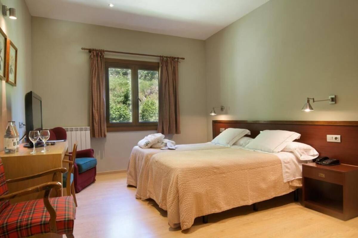 Une chambre d’hôtel avec deux lits, une table de chevet en bois, un bureau avec une chaise, une fenêtre avec des rideaux et une vue sur la verdure.