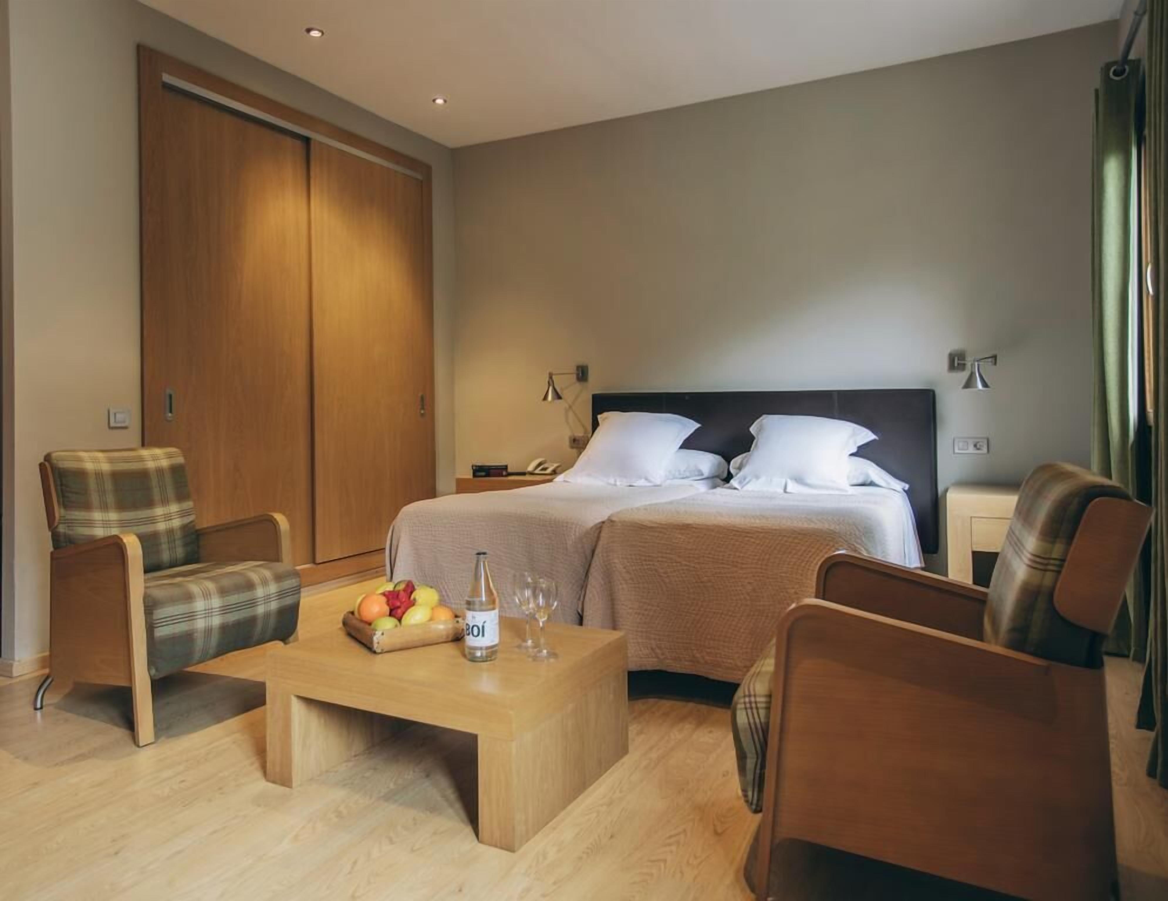 A hotel room with two beds, a wooden wardrobe, a coffee table with fruit and a bottle, and two armchairs.
