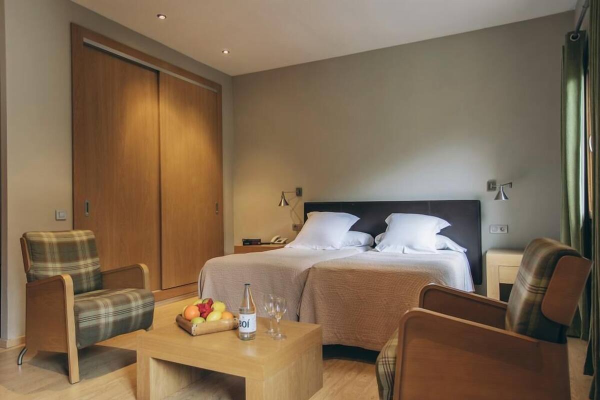 Une chambre d’hôtel avec un lit, deux chaises, une table basse en bois sur laquelle se trouvent des fruits et une bouteille, et une armoire.