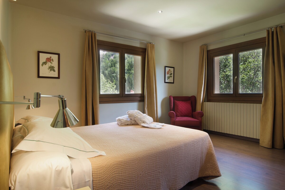 Une chambre d’hôtel avec un grand lit, une chaise, deux fenêtres avec des rideaux et un tableau au mur.