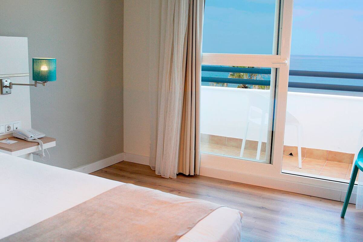 A hotel room with a bed, a bedside telephone, a nightstand, a window with curtains, and a view of the sea.