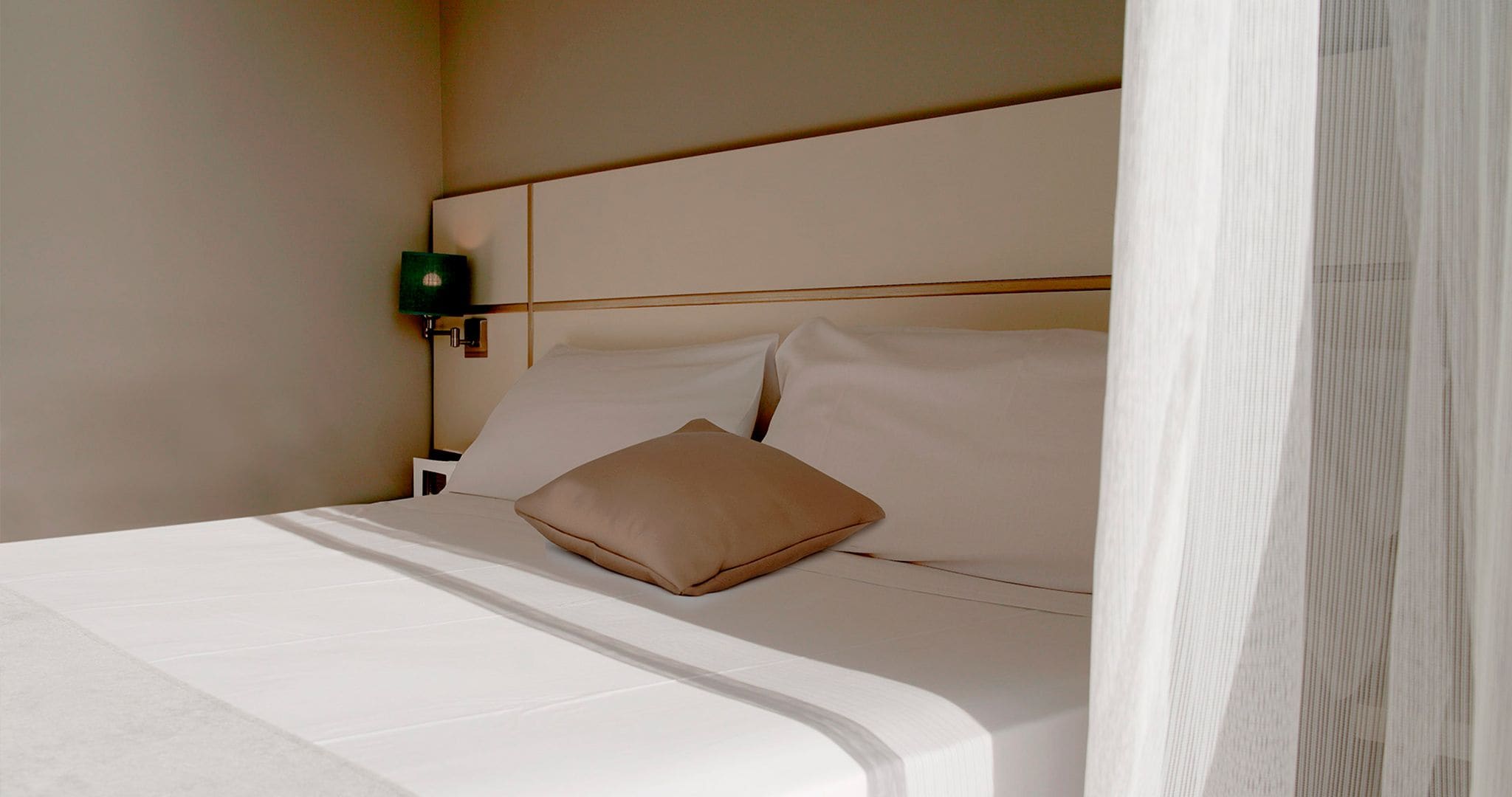 A neatly made bed with white linens and a single tan pillow, a bedside table with a green lamp, and a plain wall.