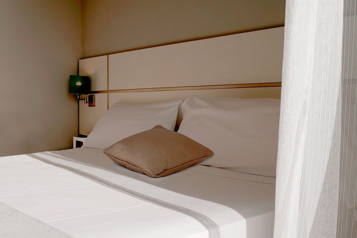 A neatly made bed with white linens and a single tan pillow, a bedside table with a green lamp, and a plain wall.