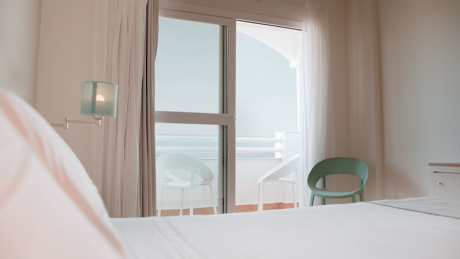 A hotel room with a bed, a chair, and a view of the sea through a sliding door.