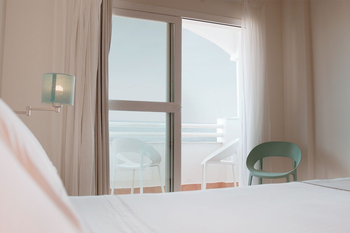 A hotel room with a bed, a chair, and a view of the sea through a sliding door.