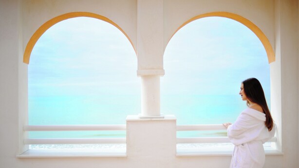 Uma pessoa vestida com um robe branco parada perto de uma janela com vista para o mar.