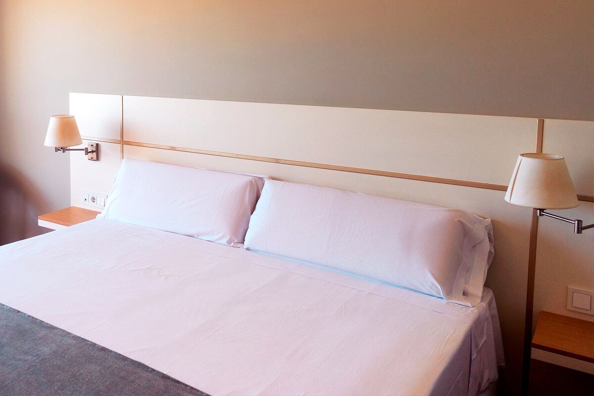 A hotel room with a large bed, two bedside lamps, a nightstand, a teal chair, and a curtain.